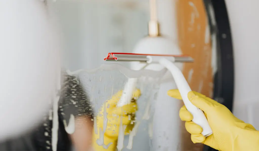man wearing yellow rubber gloves wiping water off from mirror using a squeegee