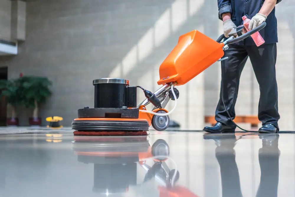 A person operates an orange floor buffer, polishing a shiny surface in a spacious, well-lit interior space